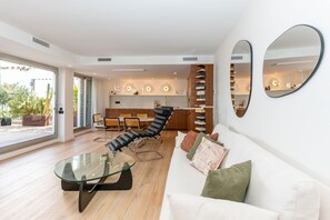 A modern living room with a glass coffee table, a leather lounge chair, a white sofa with cushions, and a kitchen in the background. - Casa Del Mar - Three Bedroom Apartment, Sleeps 6 (Sant Pere de Ribes)