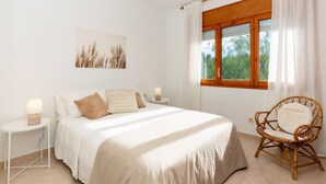 Un dormitorio con cama, mesitas de noche, una silla, una ventana con cortinas y un cuadro en la pared.