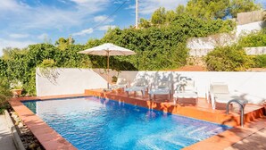 Una piscina rectangular con borde de azulejos, rodeada por un borde rojo. Hay sillas de descanso y una sombrilla blanca.