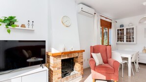 Una sala de estar con chimenea de ladrillo, televisor, un sillón rojo y un comedor con mesa y sillas blancas.