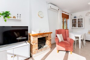 Ein Wohnzimmer mit Kamin aus Backstein, einem Fernseher, einem roten Sessel und einem Essbereich mit weißem Tisch und Stühlen.