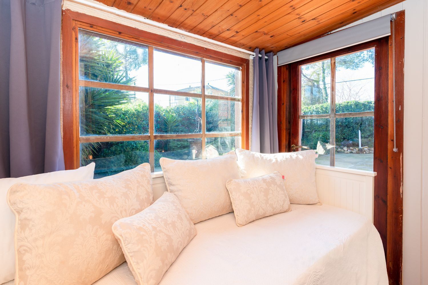 Ein Zimmer mit Holzboden, einem weißen Sofa mit gemusterten Kissen und großen Fenstern mit Vorhängen.
