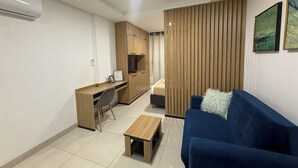 Un ambiente compatto con angolo cottura, un divano blu, un piccolo tavolo di legno e una parete divisoria con listelli verticali.