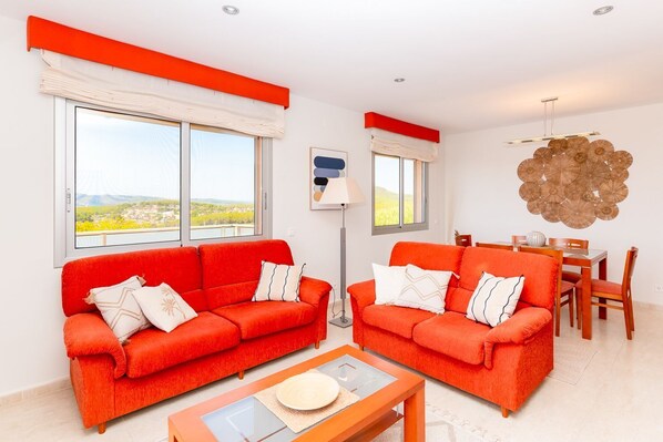 Ein Wohnzimmer mit zwei roten Sofas, einem Esstisch mit Stühlen und großen Fenstern mit Blick.