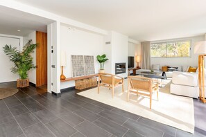 Ein modernes Wohnzimmer mit dunklen Fliesen, einem weißen Sofa, einem Essbereich mit Korbmöbeln und einem großen Fenster mit Vorhängen.