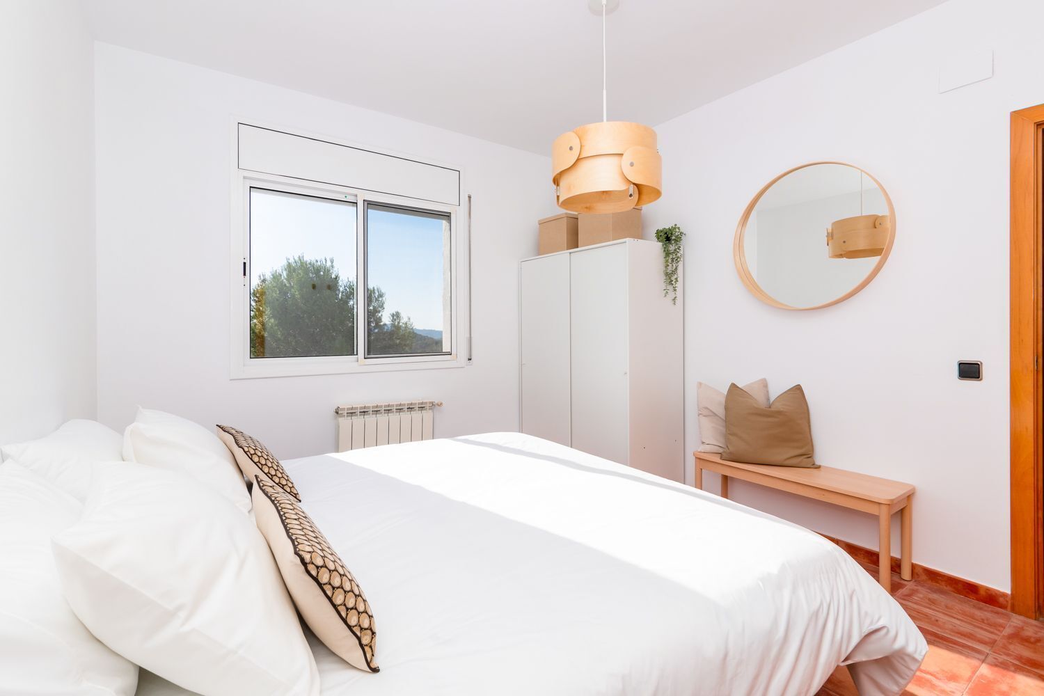 A bedroom with a bed, a window, a radiator, a bench, a mirror, and a hanging light fixture.