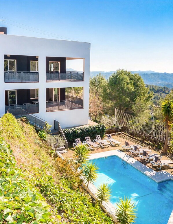 A modern two-story building with balconies, a swimming pool, and a view of the surrounding landscape.