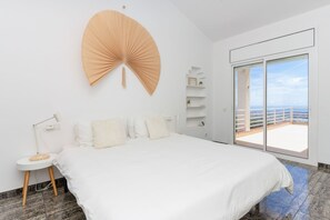 Un dormitorio con cama, mesita de noche, lámpara, un ventilador grande y vista al mar a través de una puerta corrediza.
