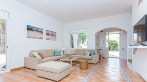 Un amplio salón con un conjunto de sofás, una mesa de centro y un televisor. Hay dos ventanas con vistas a zonas verdes.