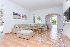 Un amplio salón con un conjunto de sofás, una mesa de centro y un televisor. Hay dos ventanas con vistas a zonas verdes.