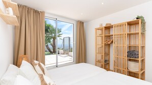 Un dormitorio con una cama grande, un armario de madera y un ventanal corredero que da a un balcón con vistas a palmeras y un edificio.