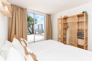 A bedroom with a large bed, a wooden wardrobe, and a sliding glass door leading to a balcony with a view of palm trees and a building. - Villa California I - Seven Bedroom Villa, Sleeps 14 (Califòrnia)