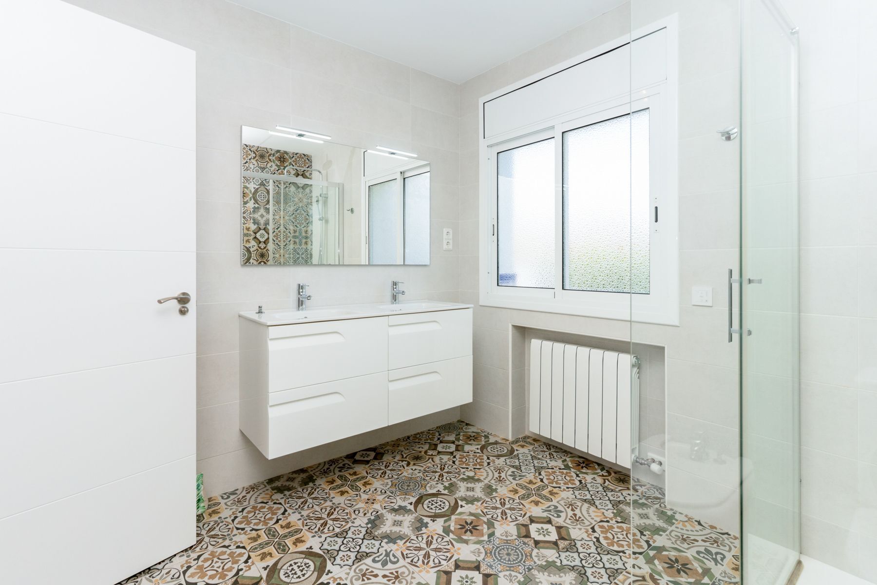 A modern bathroom with a white vanity, a large mirror, a window, and patterned floor tiles.