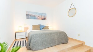 Un dormitorio con cama, mesitas de noche, lámpara, un cuadro y un espejo circular en la pared.