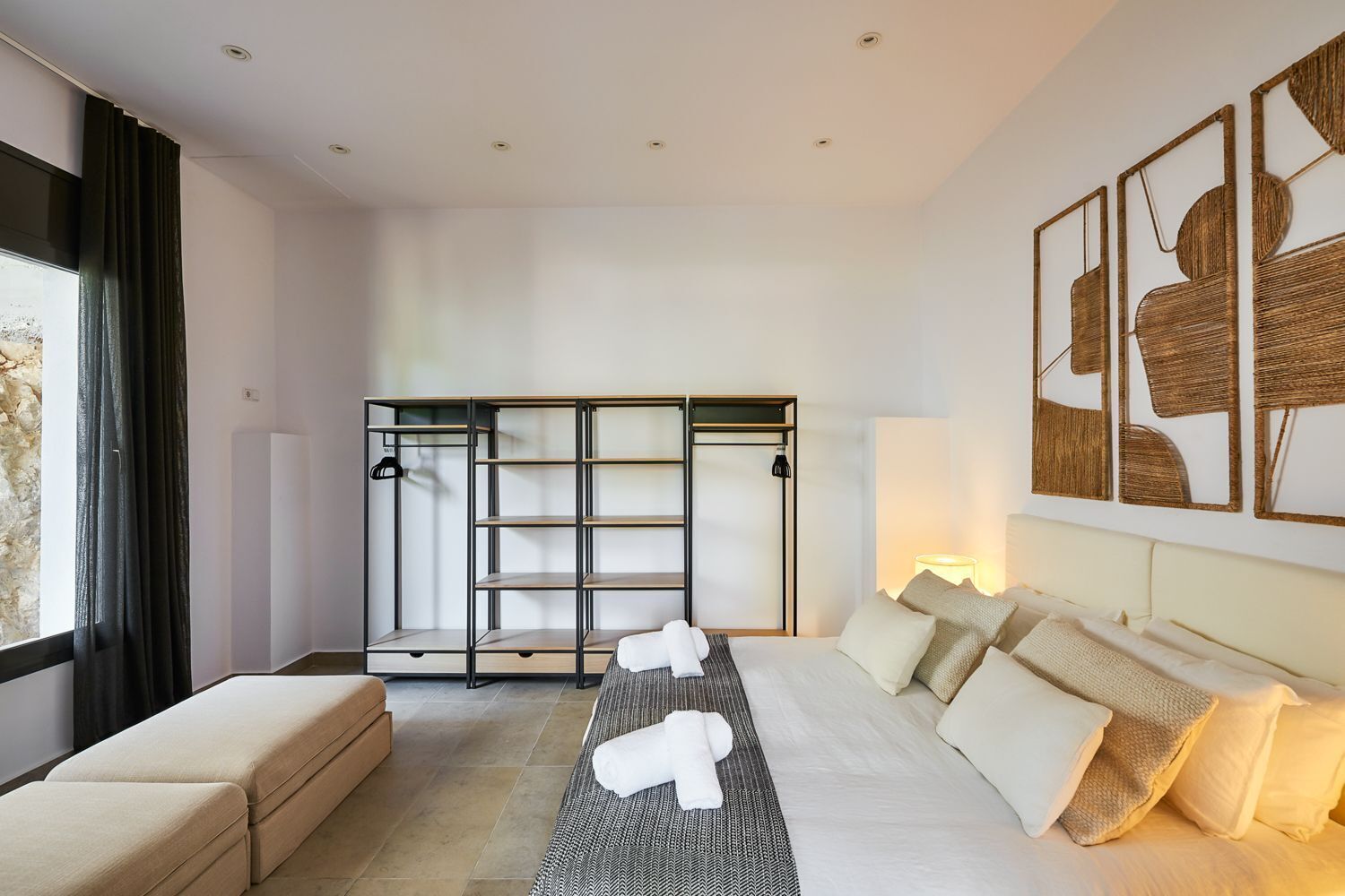 A modern hotel room with a large bed, a sofa, a built-in wardrobe, and a large window with curtains.