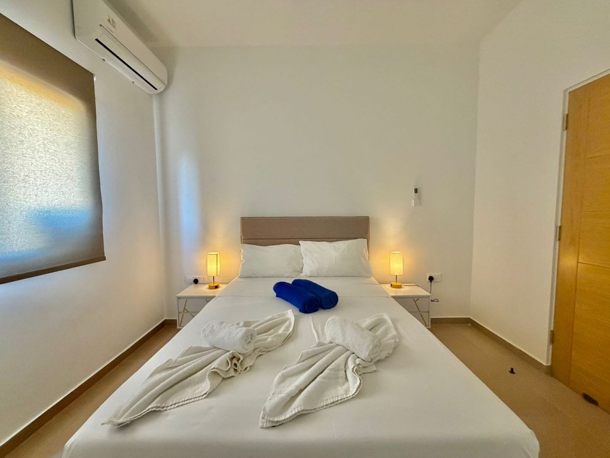 A hotel room with a bed, two bedside lamps, an air conditioning unit, and a wooden door.