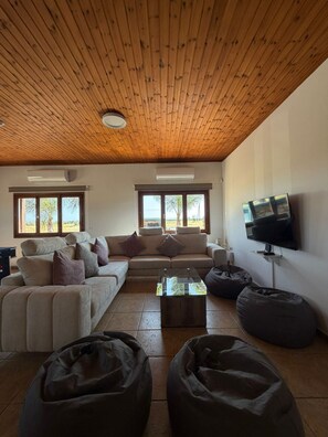 Een moderne woonkamer met een houten plafond, twee banken, een salontafel, zitzakken en een aan de muur bevestigde televisie.