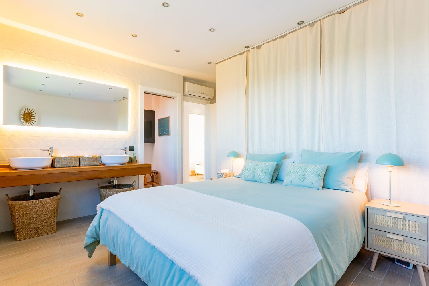 Een slaapkamer met een groot bed, nachtkastje, een badkamer met wastafel en spiegel, en een wandairconditioner.