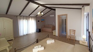 A room with a bed, a sofa, a wooden chair, a desk, a wardrobe, a shower, and a ceiling with exposed beams. - B&B Villa Sara Falconara (Butera)