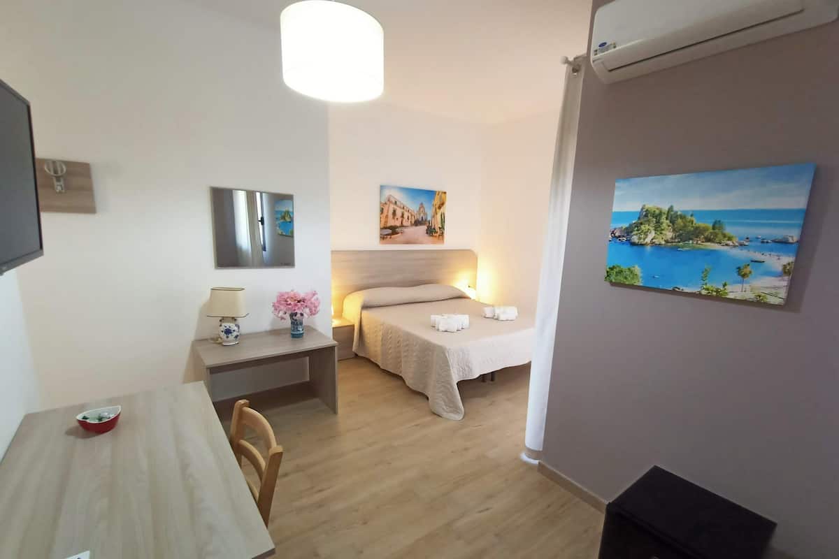 A hotel room with a bed, bedside table, lamp, and a painting of a coastal scene.