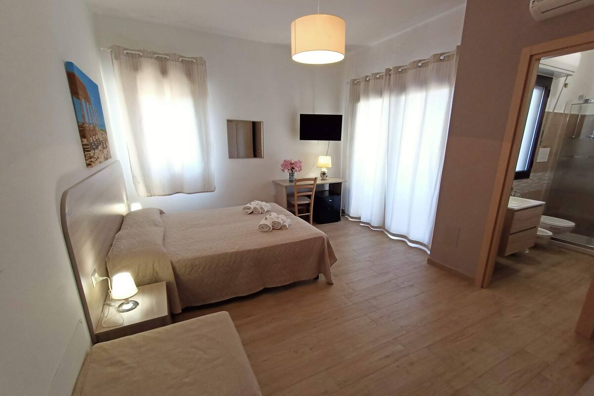 A hotel room with a bed, bedside table, lamp, desk with chair, TV, and a bathroom visible through an open door.