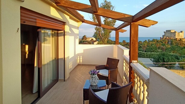 A balcony with a table and chairs, a sliding door, and a view of the sea and a castle in the distance. - B&B Villa Sara Falconara (Butera)