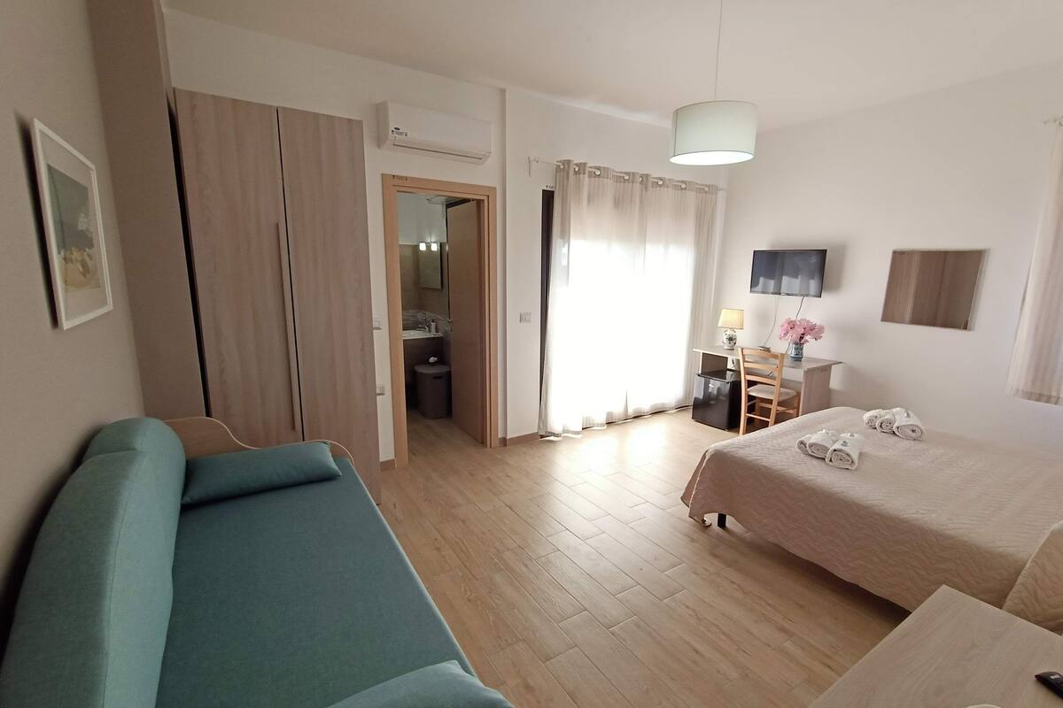A hotel room with a bed, a desk, a chair, a TV, and a bathroom visible through an open door.