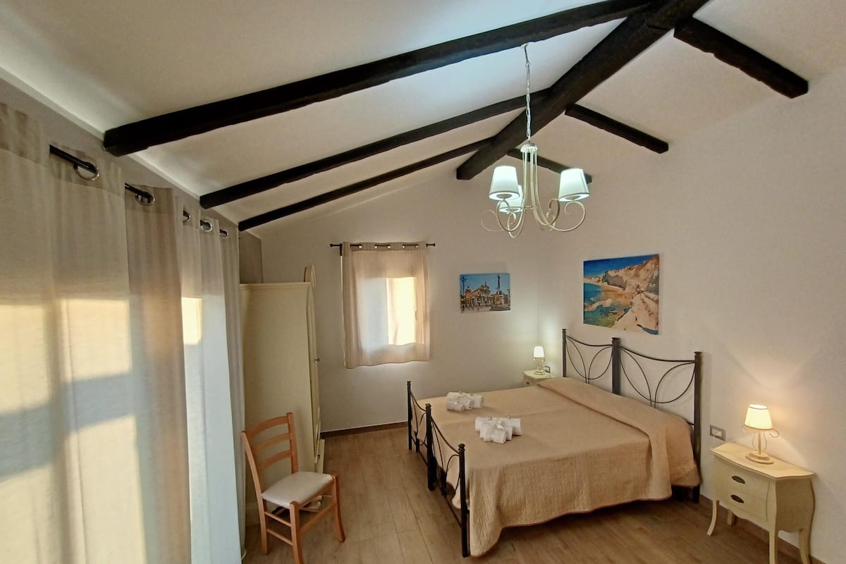 A bedroom with a bed, a chair, a nightstand, a chandelier, and two wall-mounted pictures.