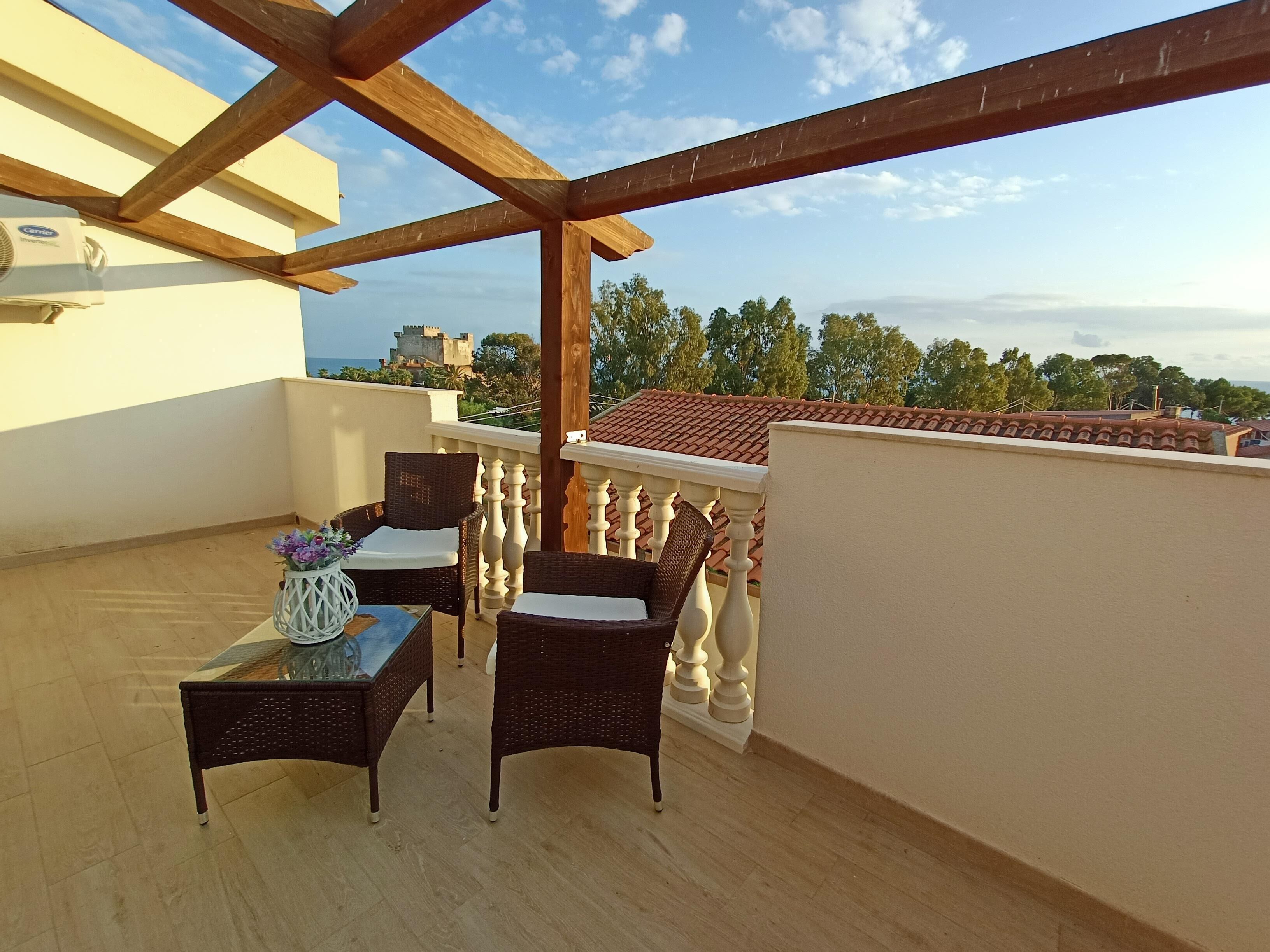 Ein Balkon mit Korbmöbeln, einem Glastisch und Blick auf ein Schloss und Bäume.