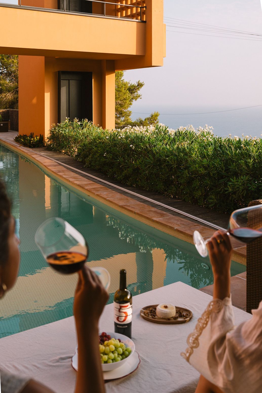 A poolside dining area with a view of the sea, a bottle of wine, and a plate of grapes.