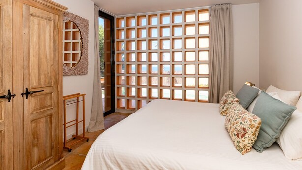 Ein Schlafzimmer mit einem Bett, einem Holzschrank, einem Fenster mit Gittermuster und Blick ins Freie.
