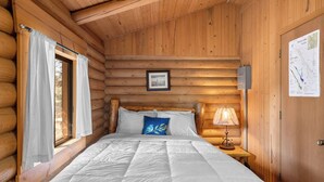A log cabin bedroom with a bed, a nightstand, a lamp, a framed picture, and a map on the wall. - New! Birdsong Log Cabin, Mountain views, firepit (Bigfork)