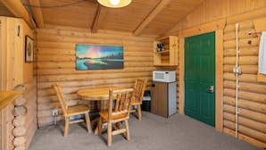 A wooden cabin interior with a dining table, chairs, a painting, a green door, and a small kitchenette. - New! Birdsong Log Cabin, Mountain views, firepit (Bigfork)