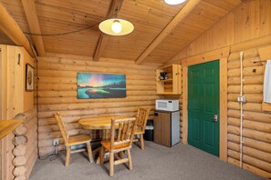 A wooden cabin interior with a dining table, chairs, a painting, a green door, and a small kitchenette. - New! Birdsong Log Cabin, Mountain views, firepit (Bigfork)