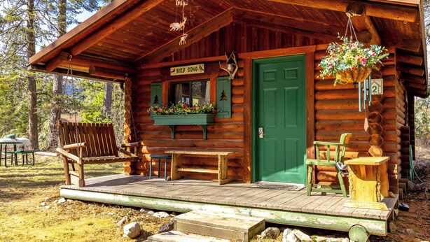 A rustic cabin with a green door, a wooden porch with benches, and a hanging flower basket. - New! Birdsong Log Cabin, Mountain views, firepit (Bigfork)