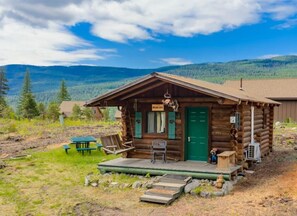 A rustic log cabin with a green door, a porch with a table and chairs, and a view of the surrounding forest and mountains. - New! Bears Den Log Cabin, Mountain views, firepit (Bigfork)