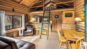 Interior de cabana de madeira com quarto em mezanino, mesa de jantar, cadeiras, sofá e escada.