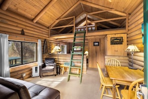 Ein Blockhaus-Interieur mit Schlafzimmer auf dem Schlafboden, einem Esstisch, Stühlen, einer Couch und einer Leitern.