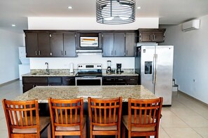 A modern kitchen with granite countertops, dark cabinets, a stainless steel refrigerator, and a central island with chairs. - Casa Postal (Puerto Peñasco)