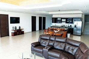 A modern living room with a leather sofa, a dining area with a wooden table and chairs, a kitchen with stainless steel appliances, and a flat-screen TV mounted on the wall. - Casa Postal (Puerto Peñasco)