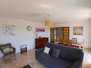 A living room with a sofa, a dining table, a chair, a small cabinet, and a wall clock. - Gîte 500m from the sea in Les Issambres - first floor (ROQUEBRUNE SUR ARGENS)