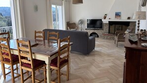 A dining area with wooden chairs and a table, a living room with a sofa and TV, a fireplace, and a balcony with a view. - Vacation Rental 8-minute walk from the ocean - top floor of a villa (ROQUEBRUNE SUR ARGENS)