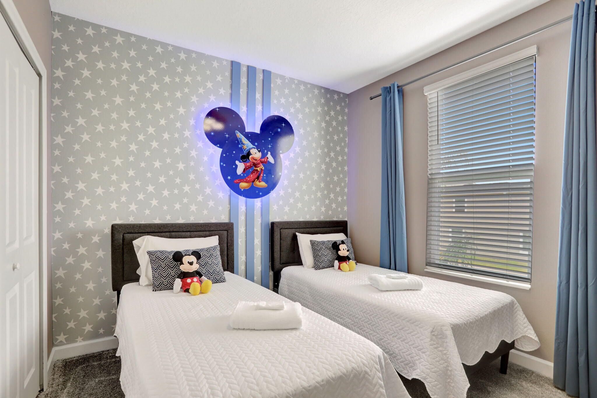 A hotel room with two beds, star-patterned wallpaper, Mickey Mouse decor, and a window with blinds.