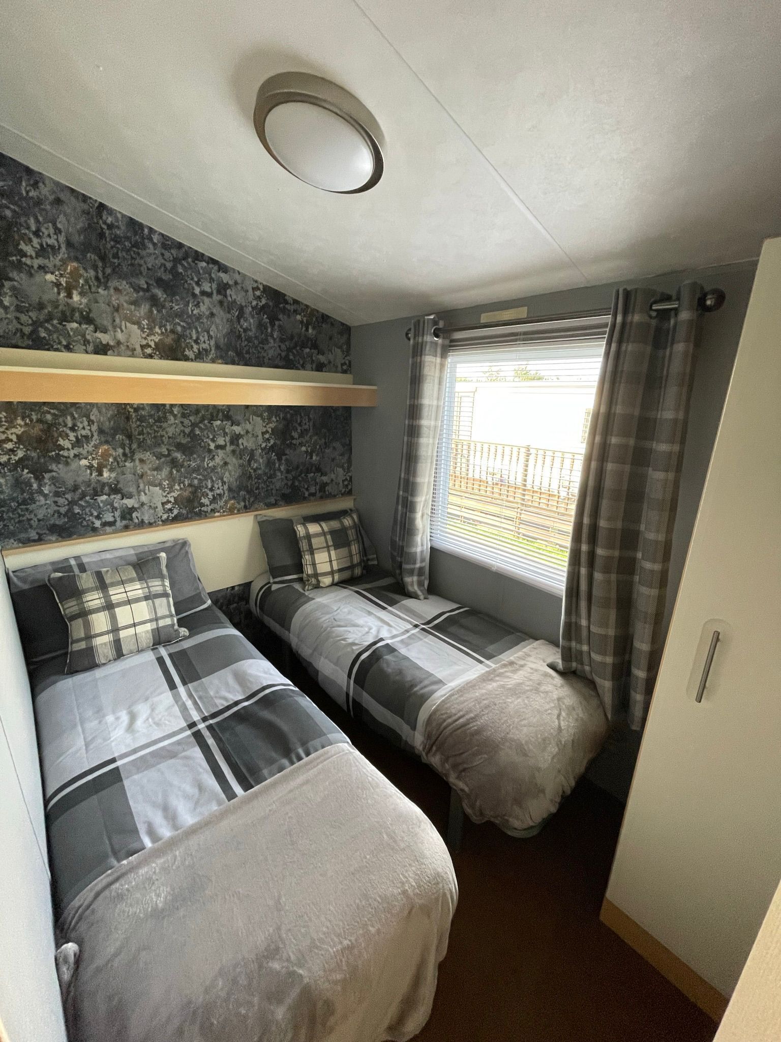A compact room with two beds, a window with curtains, a ceiling light, and a wall with a textured pattern.