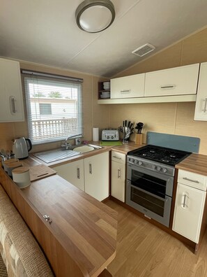 A compact kitchen with a built-in oven, a stovetop, a sink, and a window with blinds. - Golden Palm Resort, The Carnival S187 - Coastal Caravan Breaks (Chapel Saint Leonards)