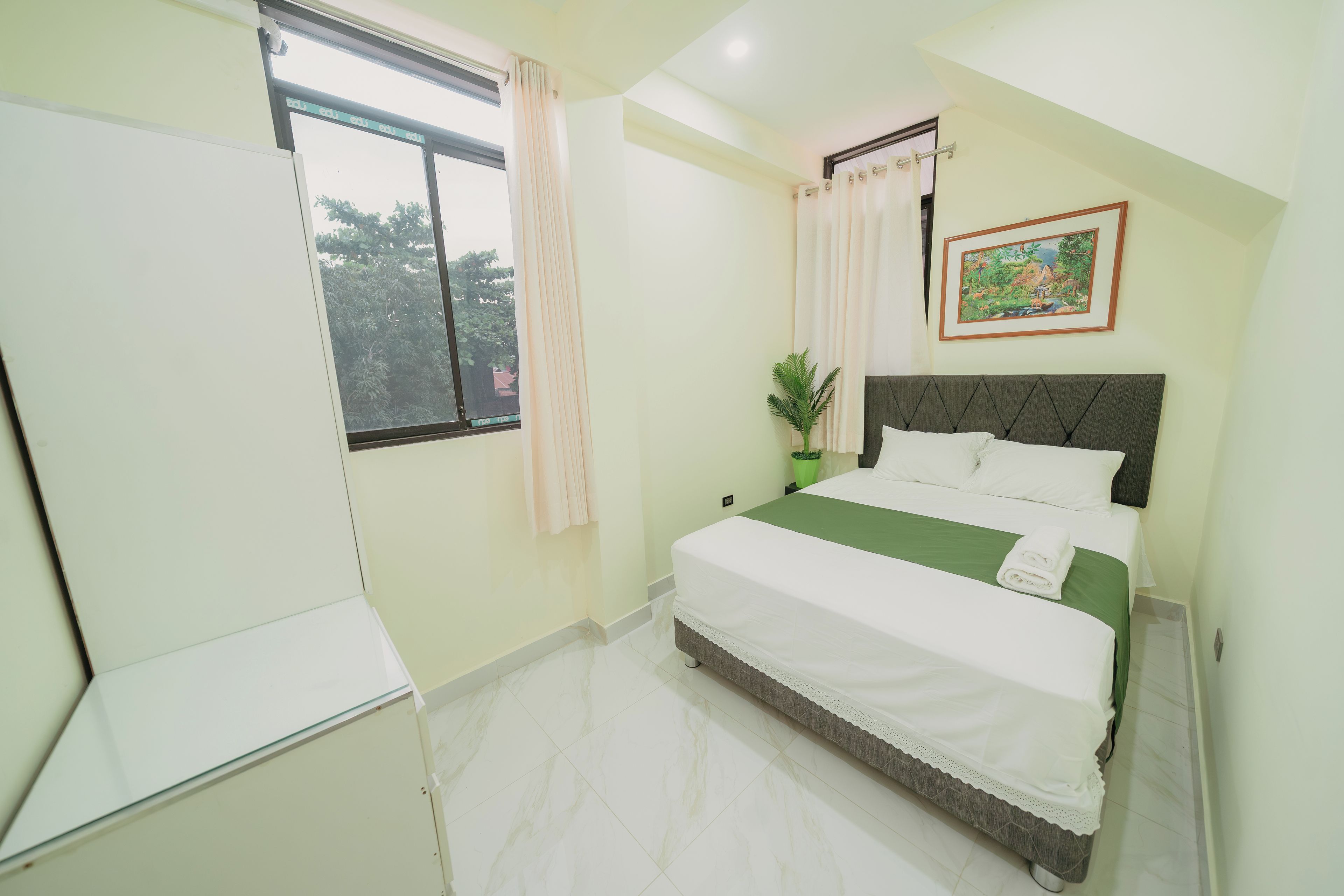 Un dormitorio con cama, cortinas en la ventana, una planta verde y un cuadro en la pared.