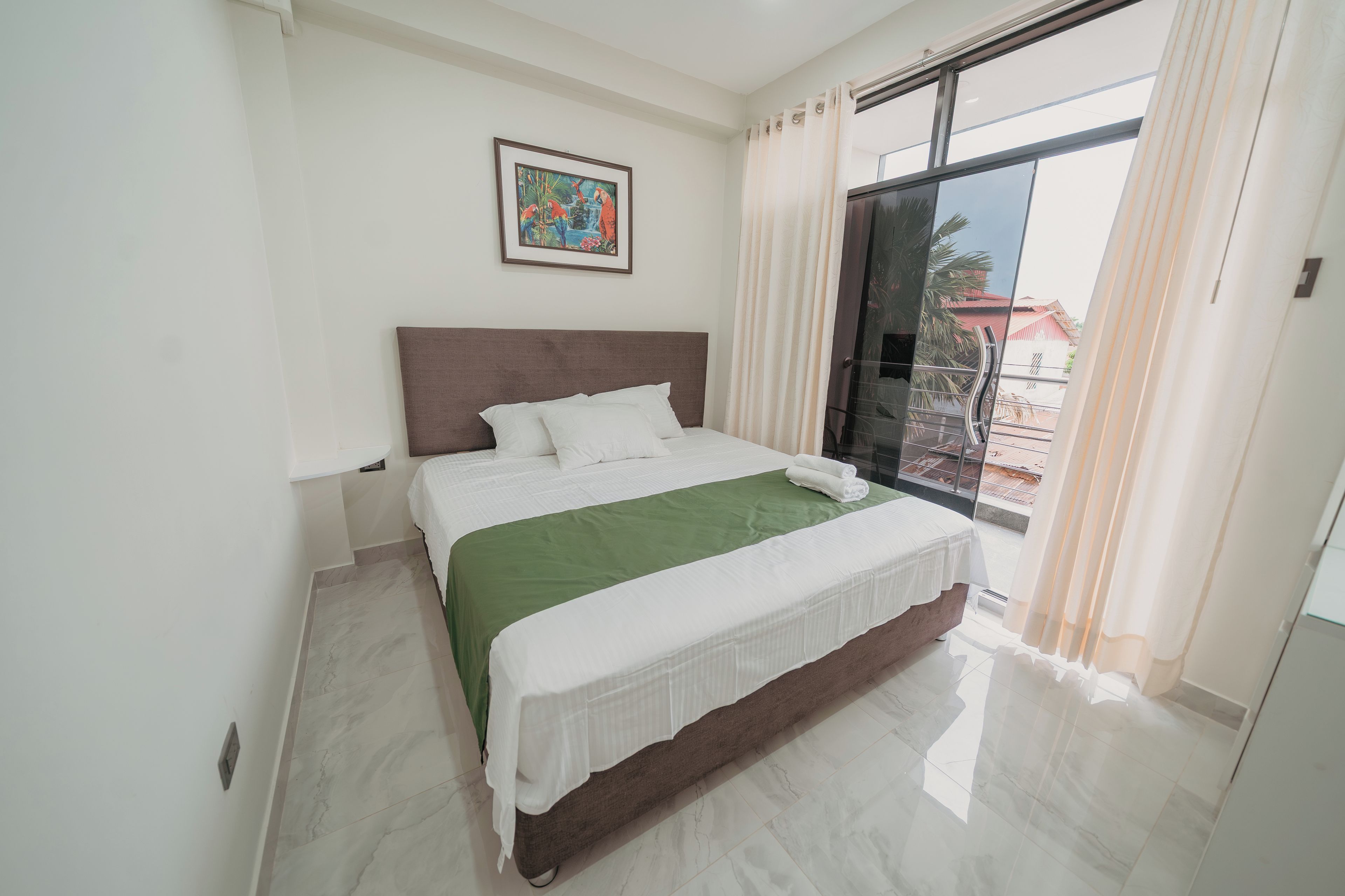 A bedroom with a bed, a green bedspread, a white headboard, a framed picture on the wall, and a view of buildings through a window.