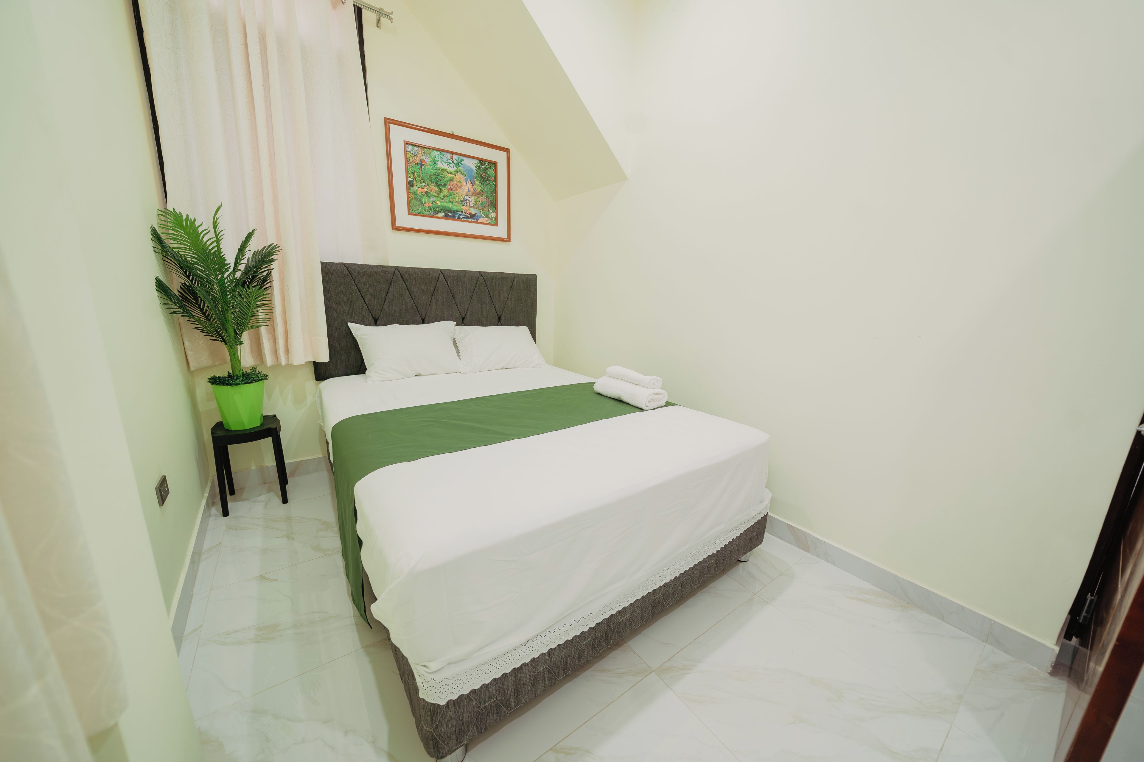 A neatly made bed with white linens, a green throw, and a decorative pillow. A potted plant is placed on a small table beside the bed.