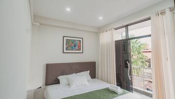 Habitación de hotel con cama, un cuadro en la pared, ventana con cortinas y puerta que da a un espacio exterior.
