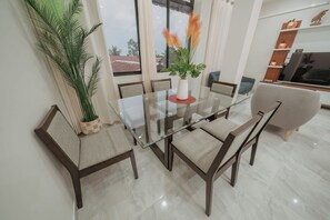 A dining area with a glass table, cushioned chairs, a potted plant, and a view through large windows.